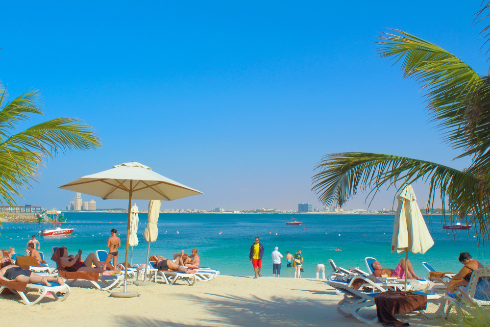 Beach Club Experience - Al Marjan Island White sand beach with loungers, cream umbrellas, palm trees, and turquoise Arabian Gulf waters at Al Marjan Island beach club lifestyle destination