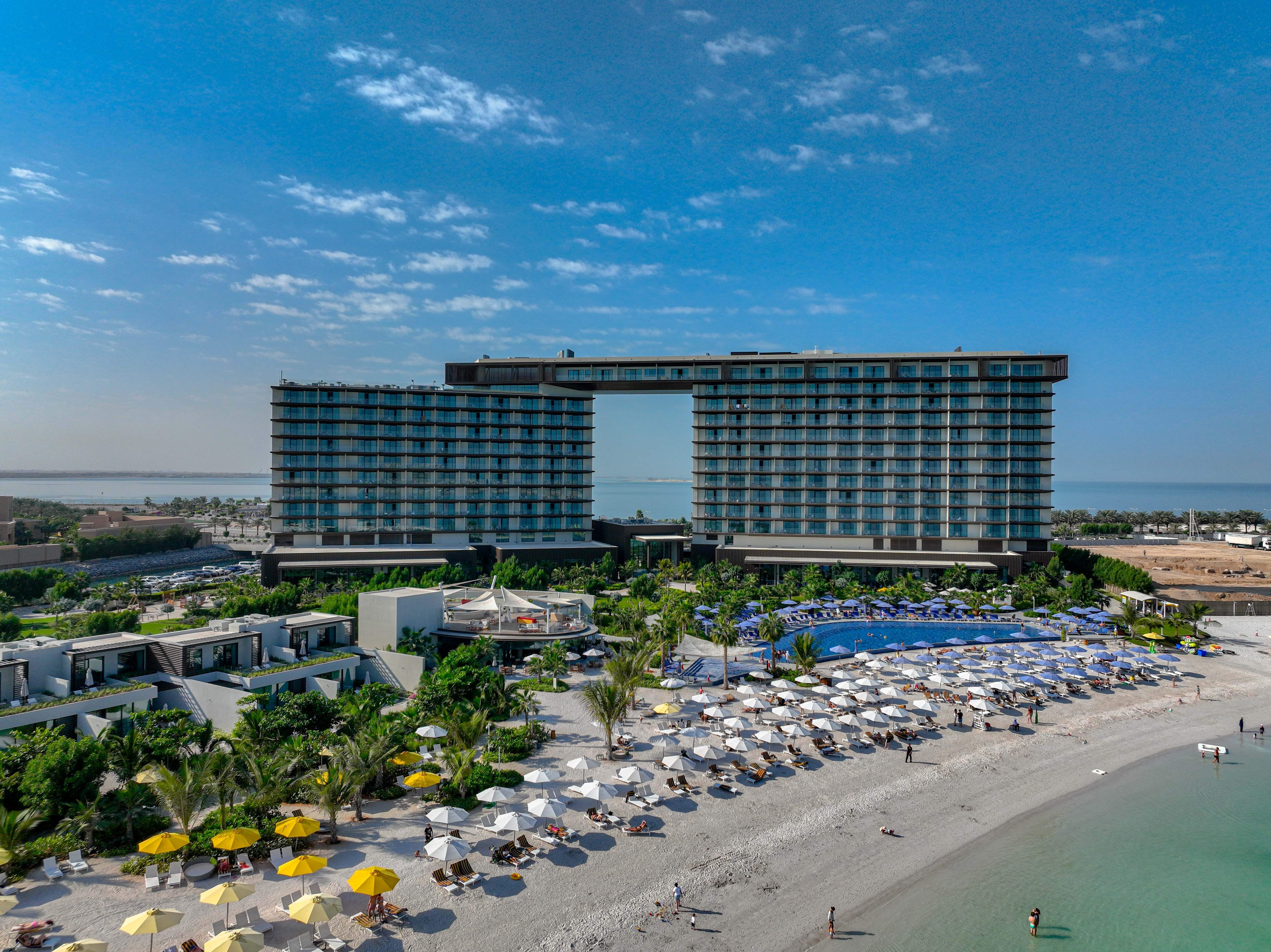 Luxury Resort Architecture - Al Marjan Island Aerial view of luxury beachfront resort with twin towers connected by sky bridge, expansive pool complex, white sand beach, and turquoise waters at Al Marjan Island